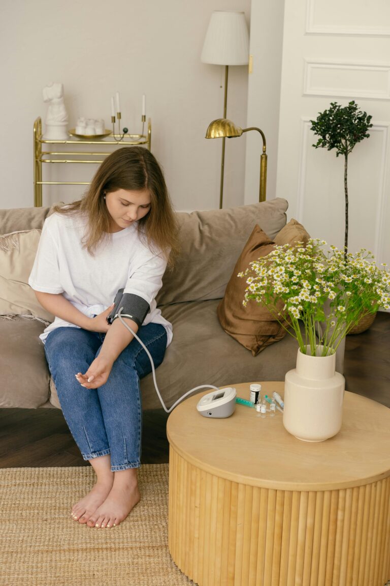 Woman sitting on couch using blood pressure monitor at home with chamomile flowers on table.